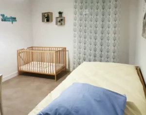 photo d'une chambre du centre départemental d'accueil mères enfants. A droite un lit une place, en fond une fenêtre avec rideaux. Sur la gauche un lit de bébé à barreaux en bois. Quelques décoration enfantines aux murs.